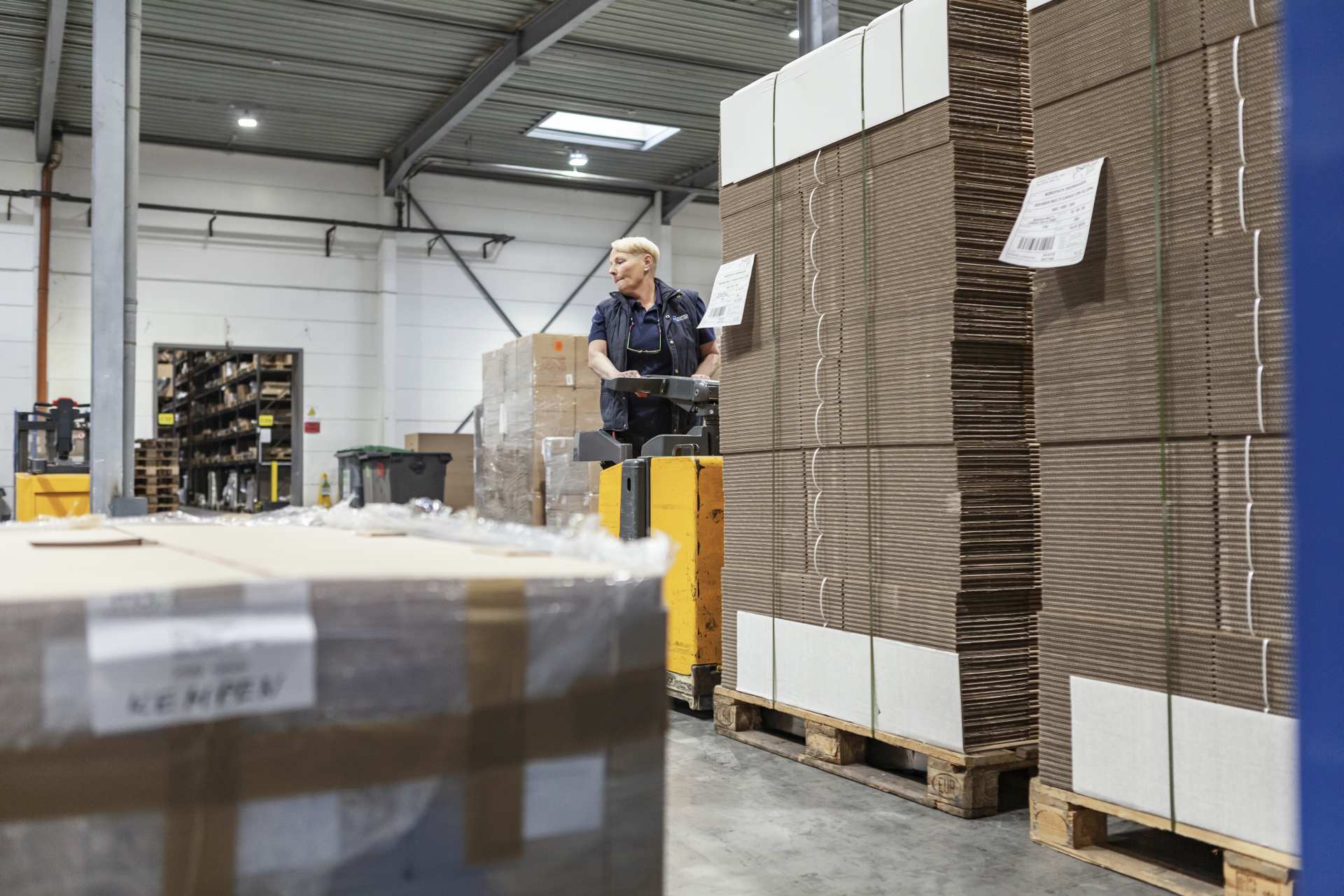 Mitarbeiter mit gelbem Hubwagen transportiert Kartonagen in Lagerhalle mit gestapelten Paketen auf Paletten