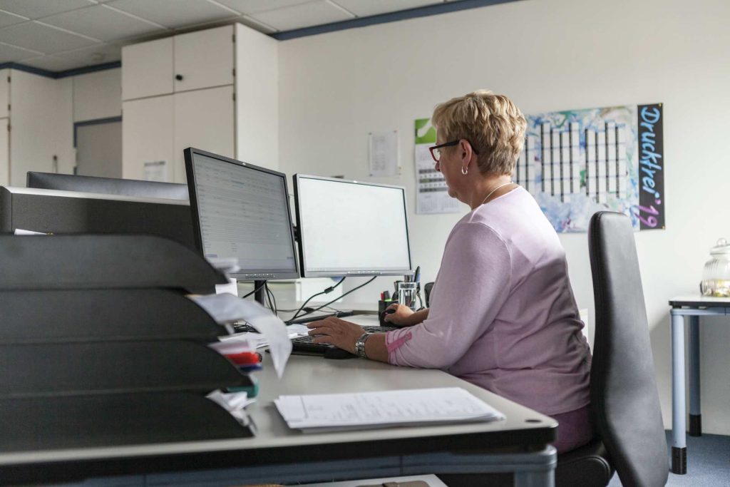 Frau mit kurzen blonden Haaren arbeitet im rosa Pullover an zwei Monitoren in modernem Büro