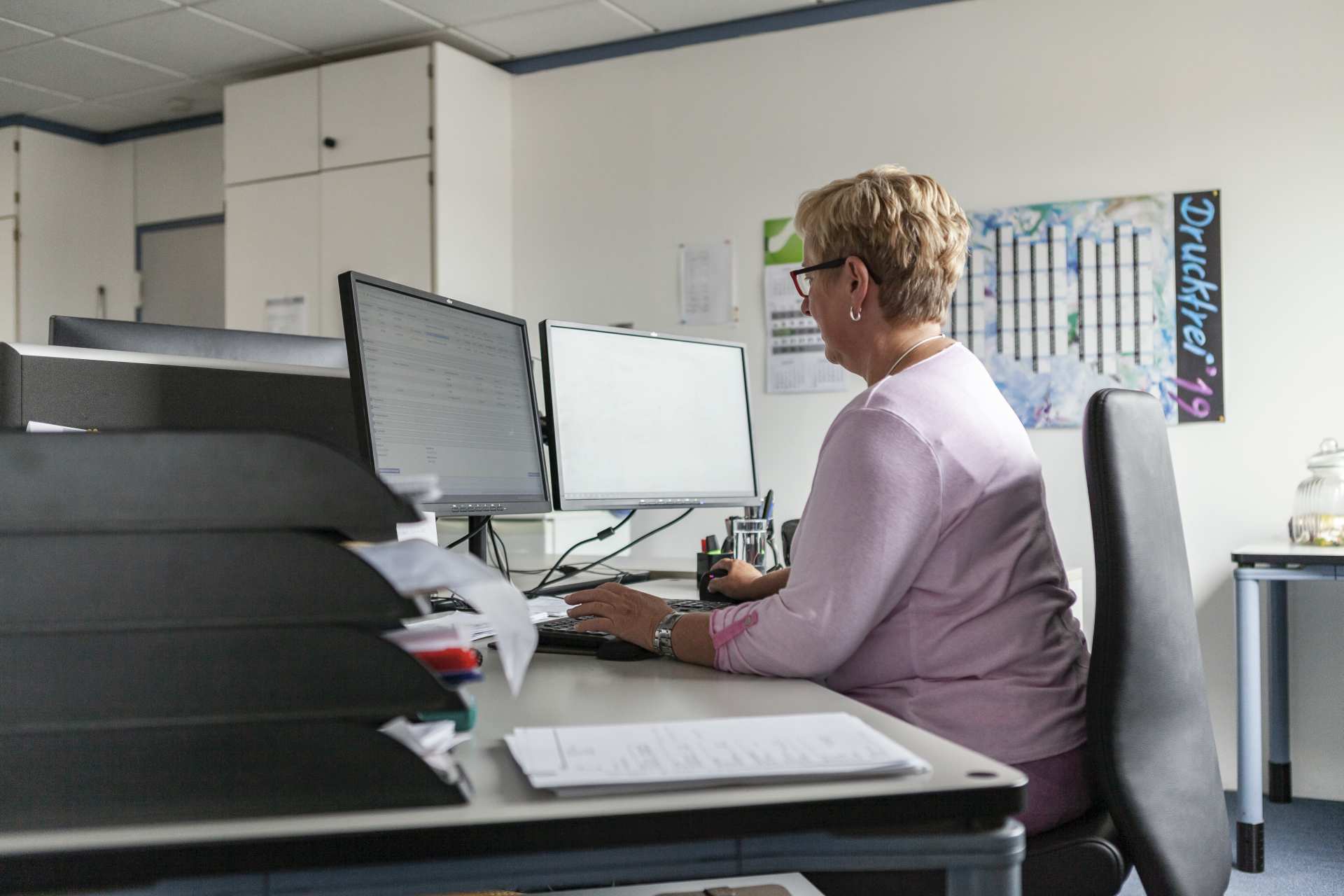 Frau mit kurzen blonden Haaren arbeitet im rosa Pullover an zwei Monitoren in modernem Büro