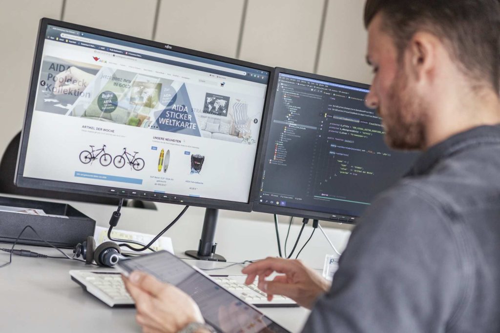 Entwickler arbeitet an zwei Monitoren mit Website-Design und Programmcode, Tablet in der Hand