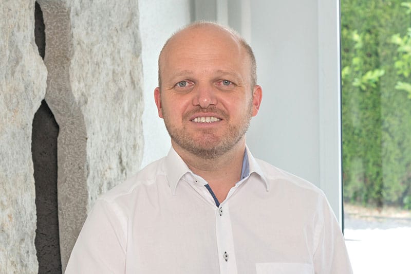 Rudolf Nopper lächelt freundlich in weißem Hemd vor moderner Betonwand und grünem Hintergrund