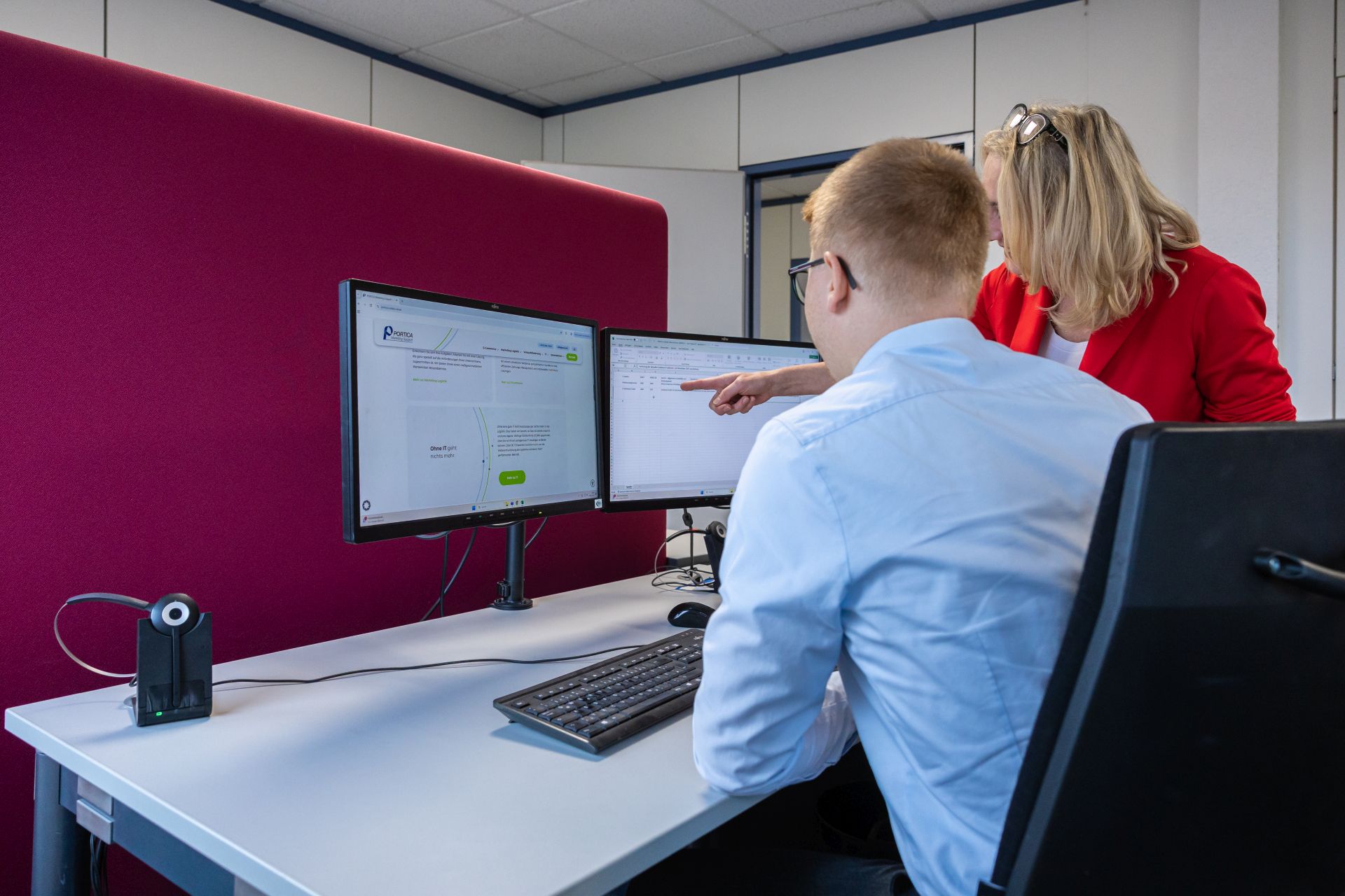 Zwei Kollegen arbeiten gemeinsam am Computer mit zwei Monitoren in modernem Büro mit roter Wand