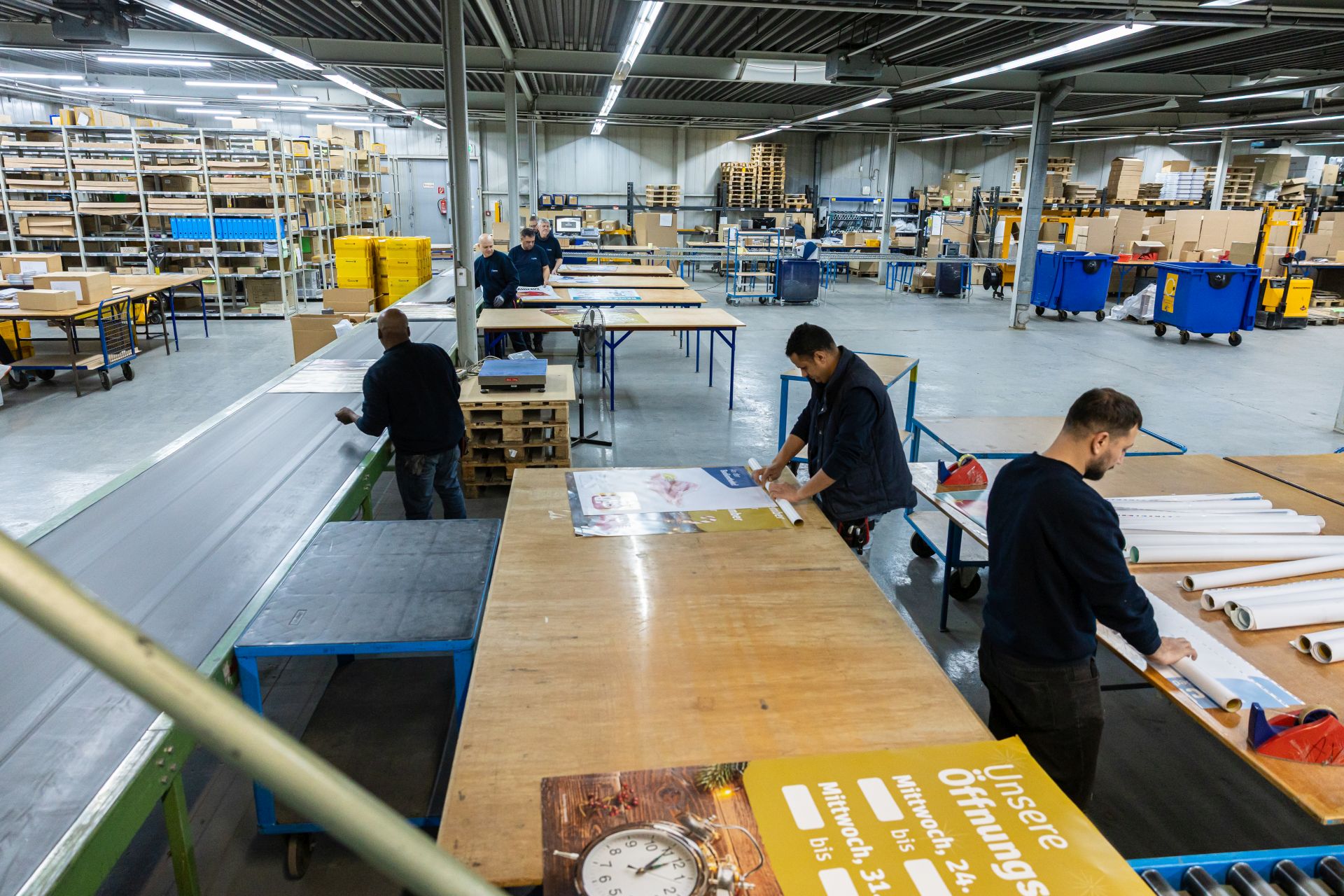 Mitarbeiter in Industriehalle verpacken Plakate und Druckerzeugnisse an Arbeitstischen zwischen Regalen und Maschinen