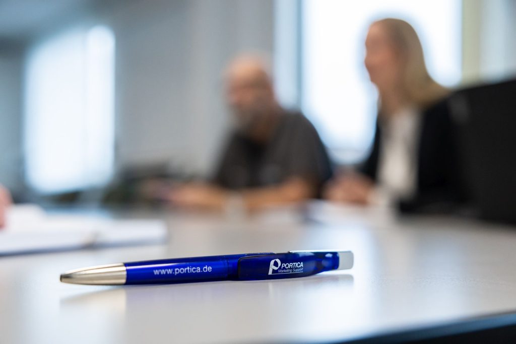 Blauer Kugelschreiber mit Portica-Logo auf weißem Tisch, im Hintergrund unscharfe Personen in Besprechungssituation