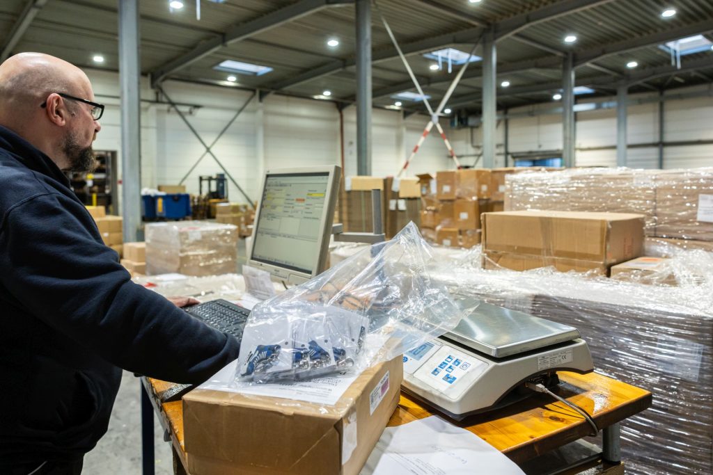 Mitarbeiter wiegt Paket auf digitaler Waage und arbeitet am Laptop in Lagerhalle mit Kartonpaletten