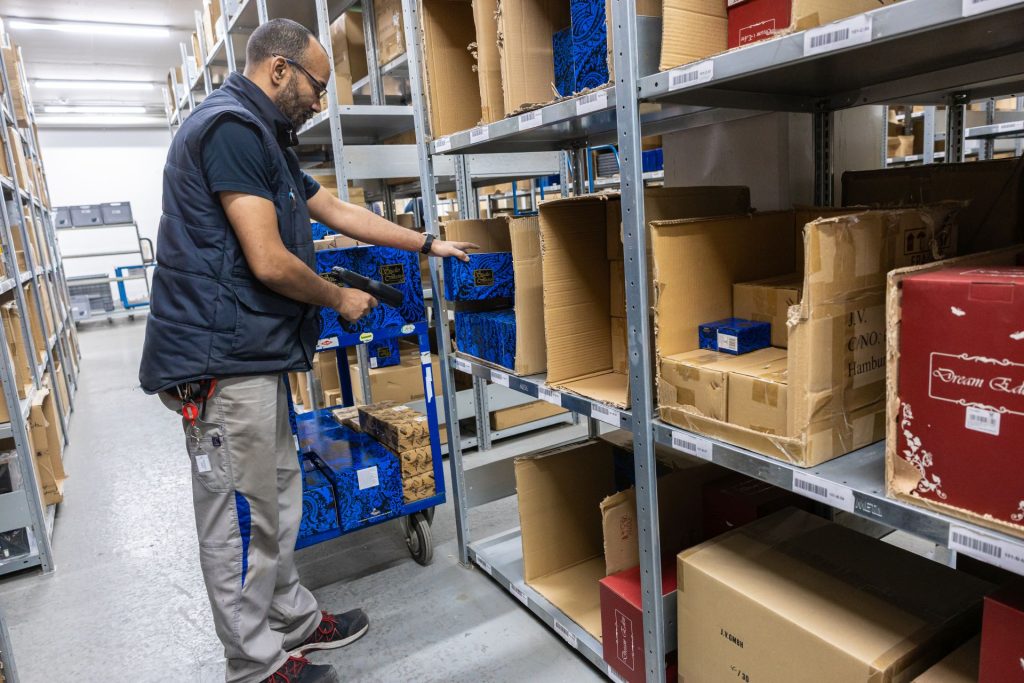 Lagerarbeiter mit Brille sortiert blaue Kartons auf Transportwagen zwischen hohen Metallregalen im Warenlager