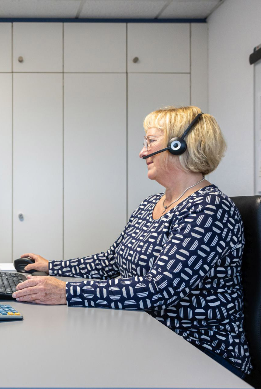 Blonde Frau mit Headset arbeitet lächelnd am Computer in modernem Büro mit grauen Schränken