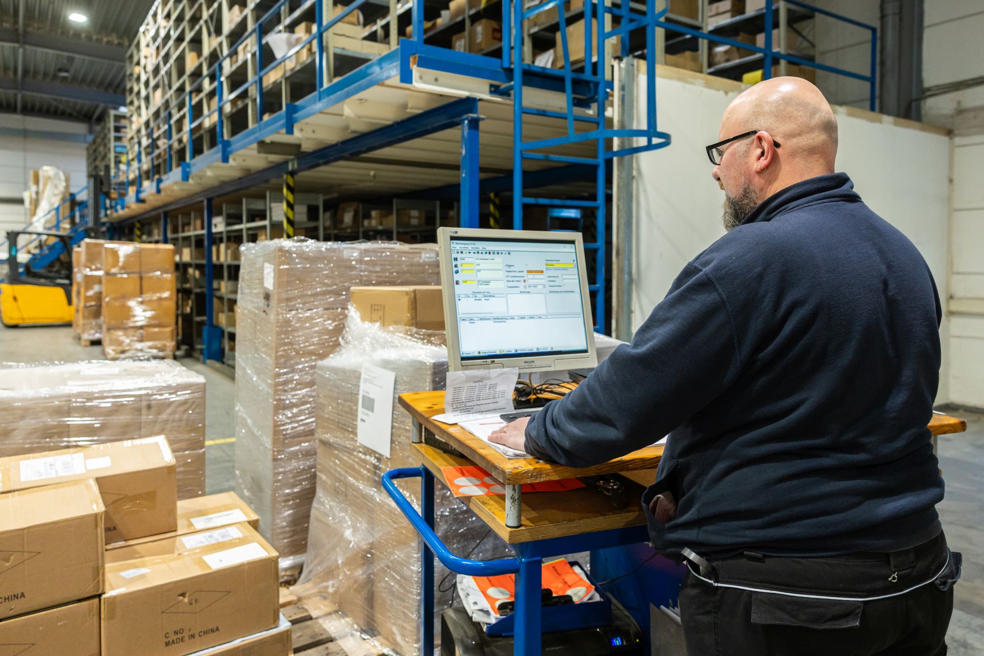 Mitarbeiter arbeitet am Laptop auf mobilem Arbeitsplatz in Lagerhalle mit blauen Regalen und verpackten Kartons